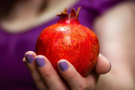 Blog author holding a pomegranate out to the viewer in her hand, with a pale purple nail polish on her fingers